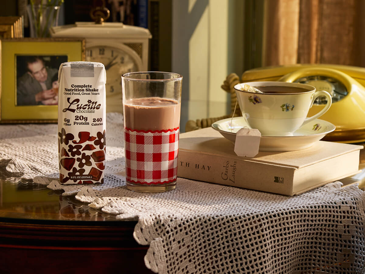 A carton of Lucille Chocolate Complete Nutrition Shake sitting on a table next to a poured glass of the shake, a book, and a cup of tea.