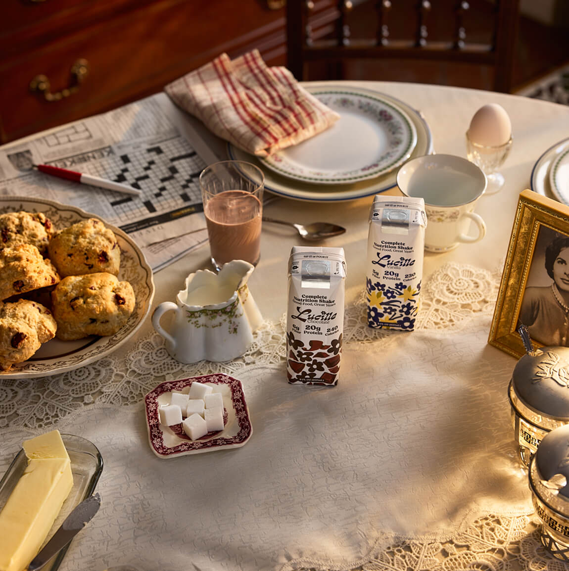 Breakfast table set with scones, a hard-boiled egg, butter, sugar, and two Lucille Complete Nutrition Shakes – Chocolate and Vanilla.